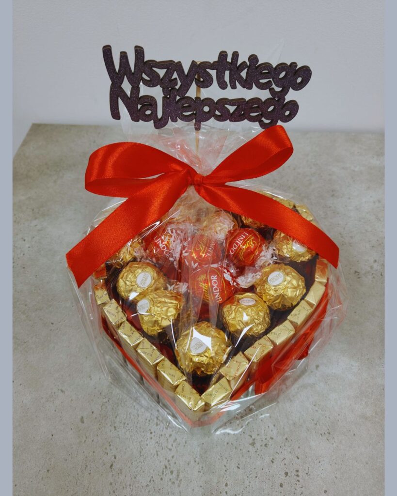 alt = "Torcik ze słodyczy w kształcie serca z pralinkami Lindt Lindor, ozdobiony czerwoną wstążką i topperem "Wszystkiego najlepszego" – idealny prezent na każdą okazję."