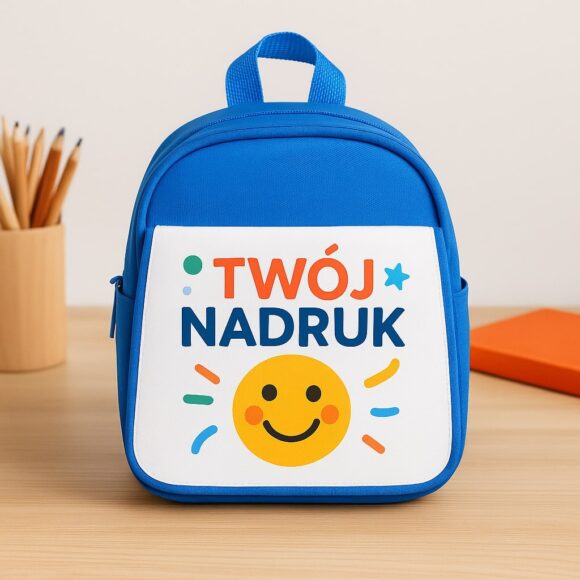 Personalizowany niebieski plecak z nadrukiem „Twój nadruk” i uśmiechniętą buźką, ustawiony na biurku obok kredek i pomarańczowego notesu – idealny do przedszkola lub na prezent