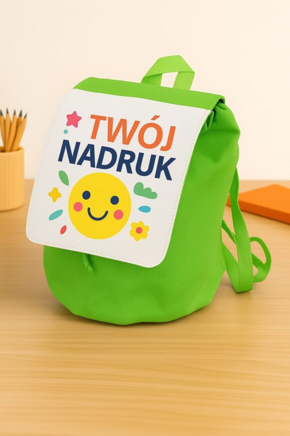 Zielony plecak dla dziecka z nadrukiem na klapie – kolorowy napis „Twój nadruk” i uśmiechnięte słońce, ustawiony na biurku obok kredek