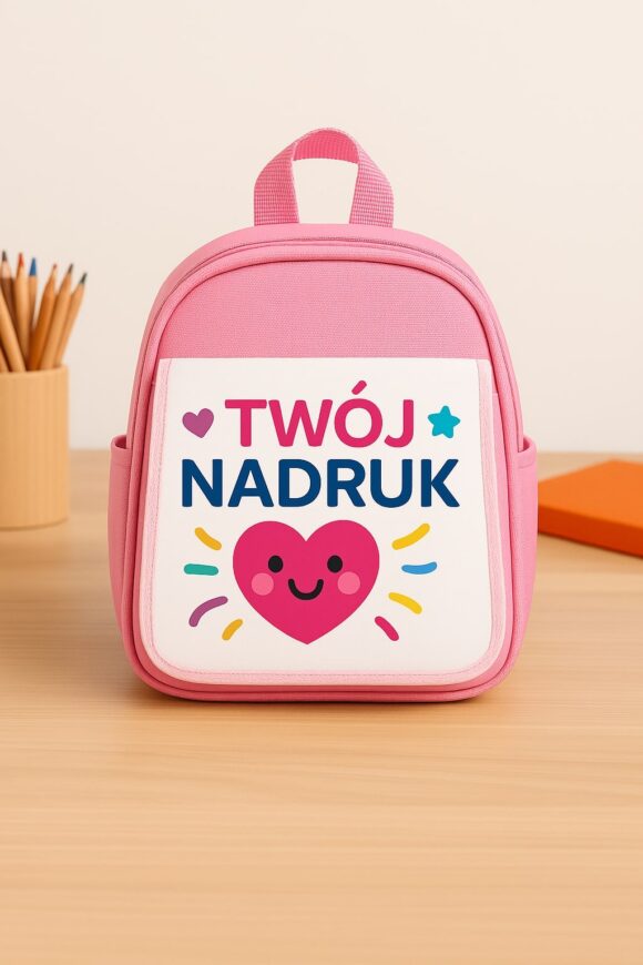 Personalizowany różowy plecak dziecięcy z nadrukiem „Twój nadruk” i rysunkiem serduszka z uśmiechem, ustawiony na drewnianym biurku obok kubka z kredkami i pomarańczowego notesu