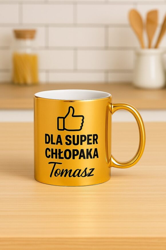Złoty kubek z grafiką kciuka i napisem „Dla super chłopaka Tomasz”, sfotografowany w kuchni na blacie obok akcesoriów kuchennych.